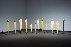 Twelve elongated diamond-shaped sculptural figures suspended from the ceiling, grouped to evoke a crowd. They are varying in size and each has a hood