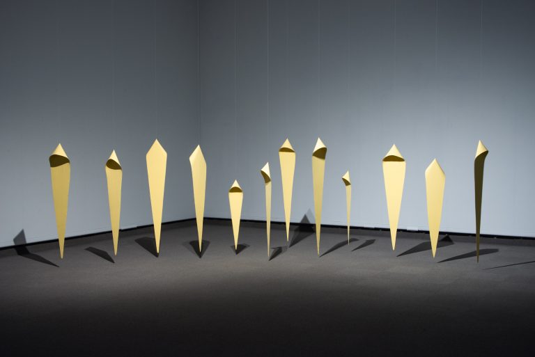 Twelve elongated diamond-shaped sculptural figures suspended from the ceiling, grouped to evoke a crowd. They are varying in size and each has a hood
