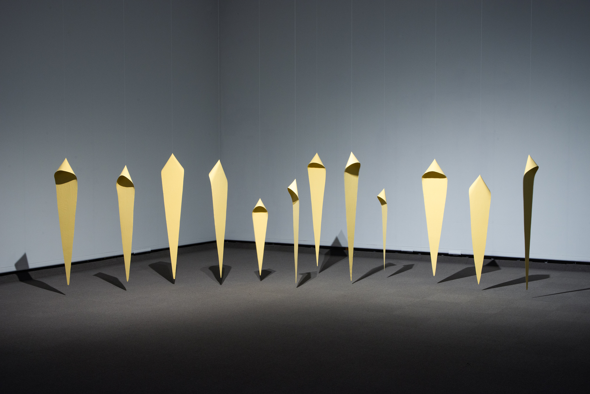 Twelve elongated diamond-shaped sculptural figures suspended from the ceiling, grouped to evoke a crowd. They are varying in size and each has a hood